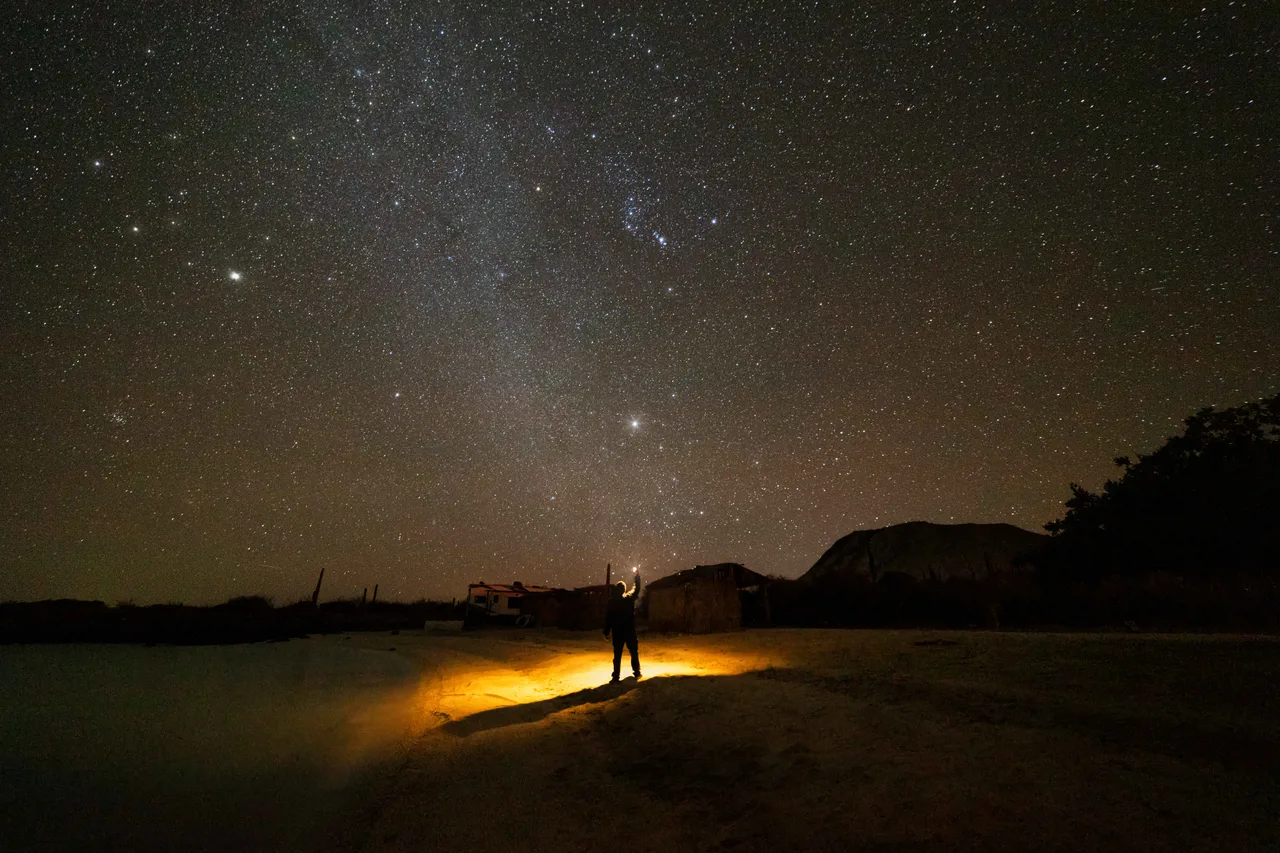 Una persona apunta una linterna hacia el cielo estrellado donde brilla Sirio, junto a una casa en el desierto.